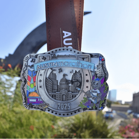 A detailed Austin Marathon 2025 belt buckle medal showcases iconic designs with embossed city landmarks, musical notes, and colorful elements like a guitar and a burger. It hangs in front of a blurred cityscape. Austin Marathon Half Marathon & 5K